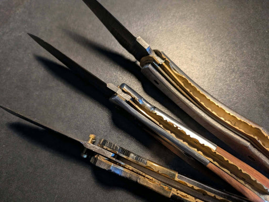 Trois couteaux pliants artisanaux à lames noires et manches métalliques et dorés, photographiés ouverts pour présenter leurs mécanismes de verrouillage et leurs finitions.